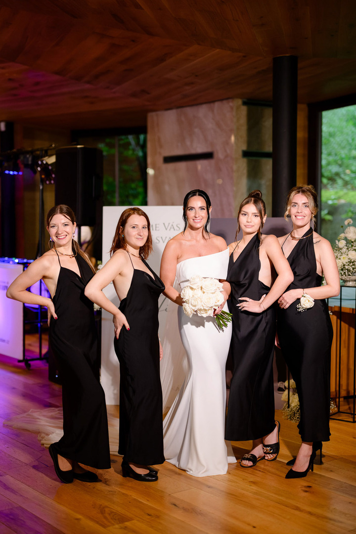 The bride in white poses with her friends and bridesmaids, who are all wearing black dresses. Only the bride holds a white bouquet.
