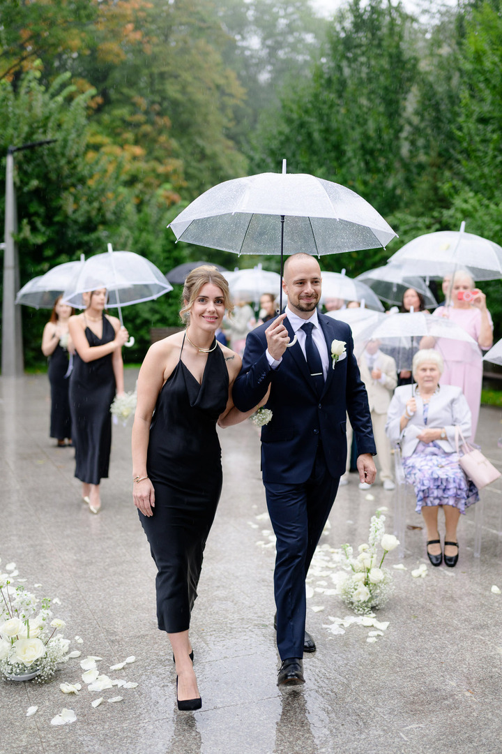 The best man and maid of honor, both in black dresses, walk together under one umbrella, approaching the wedding venue.

