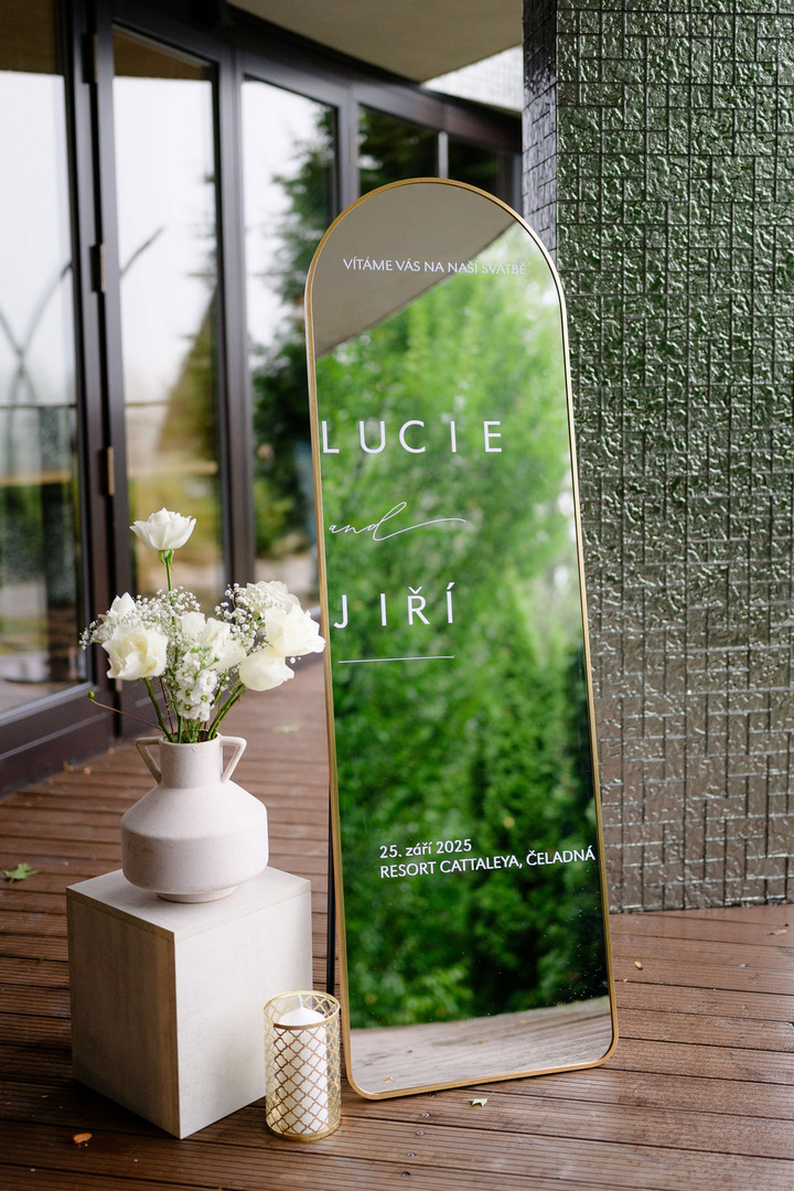 Wedding decoration – a large standing mirror with the names "Lucie and Jiří" written on it, next to a vase with white roses.
