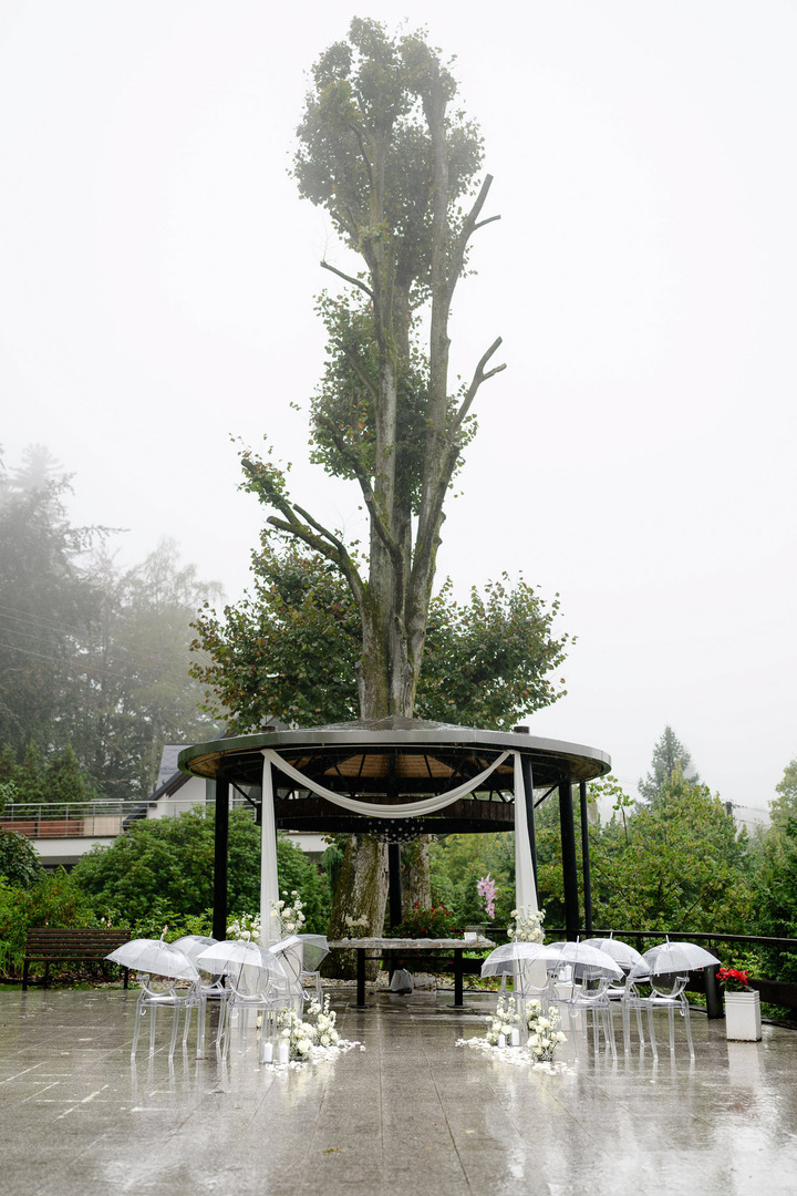 The decorated wedding gazebo in the garden of Villa Cattaleya, with mist in the background, a tall tree, and transparent umbrellas placed on the chairs.
