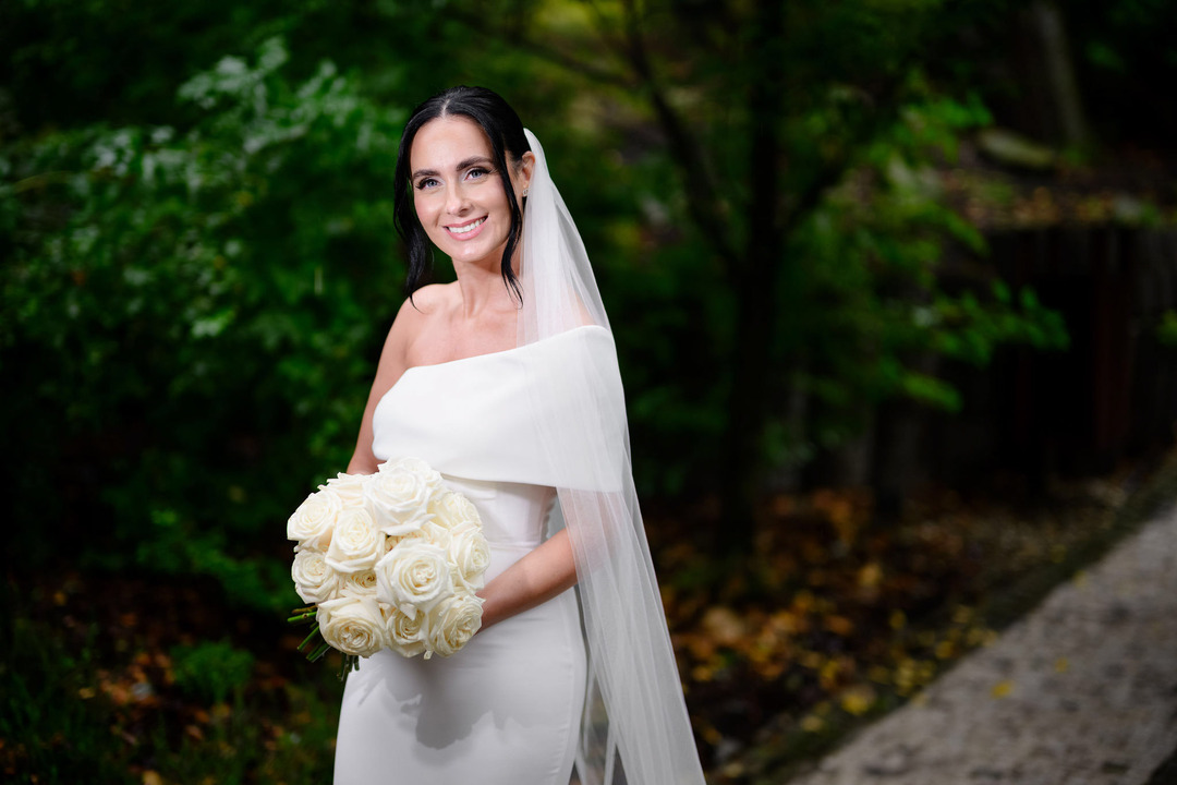 The bride poses alone in the garden, holding a bouquet of white roses and smiling at the camera.

