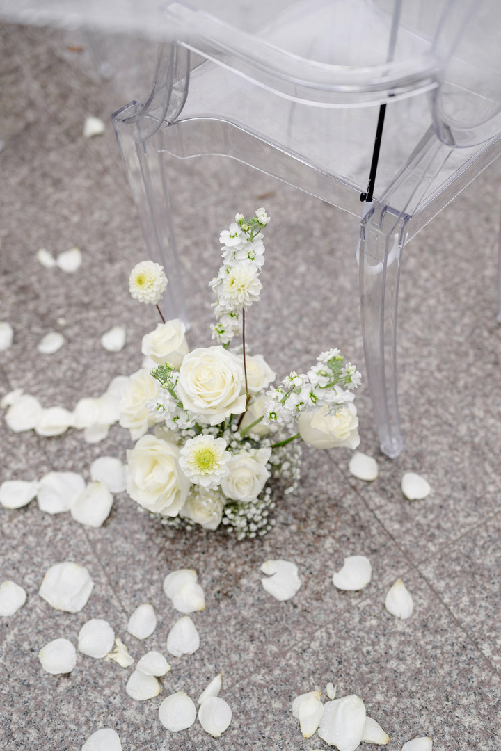 White rose arrangements placed by a chair at the wedding venue, with white petals scattered on the ground.
