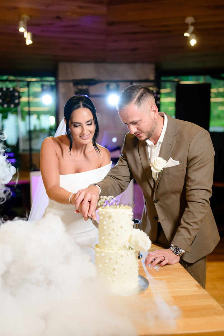 The newlyweds cut their cake, with the groom's hand on the bride's. Together, they slice the cake and smile at each other.
