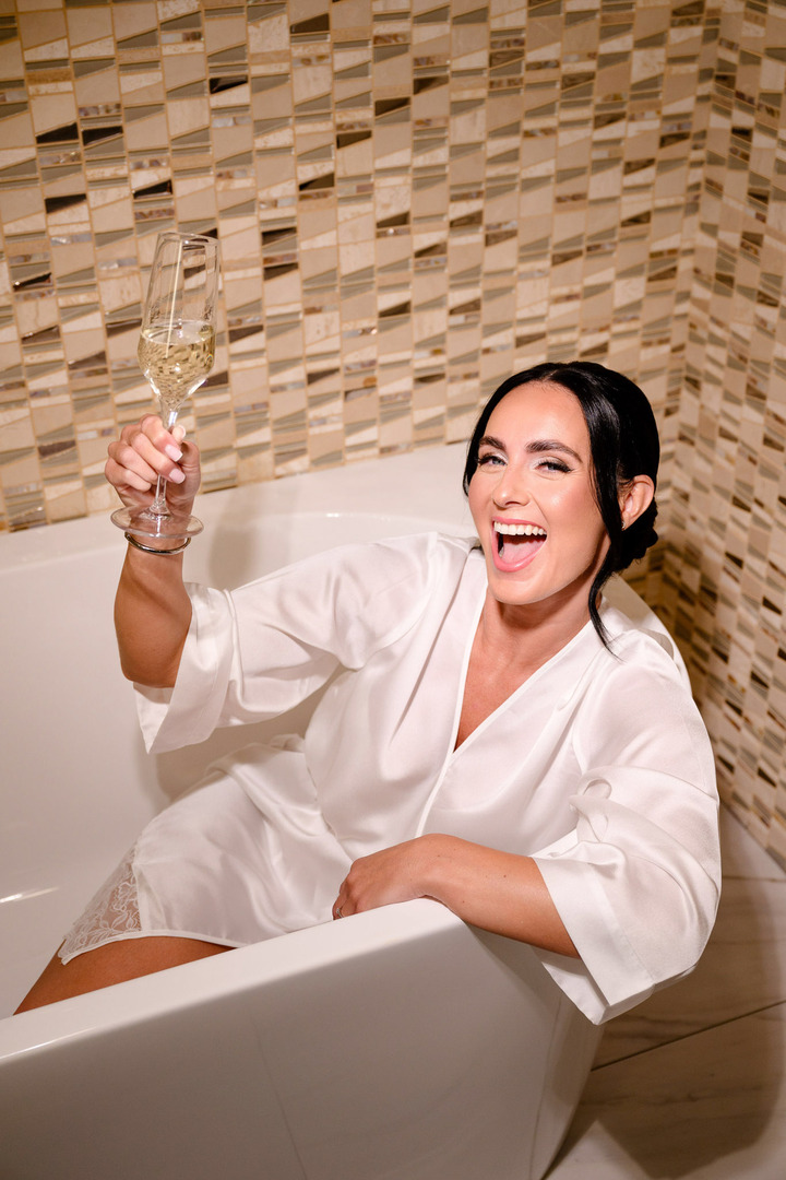 Bride sitting in a bathtub wearing her white robe, raising her champagne and laughing cheerfully.
