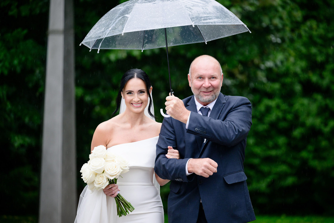 The bride approaches the altar, arm-in-arm with her father, who holds an umbrella over them as it rains.
