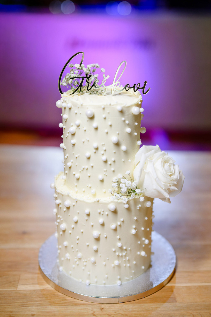 A white two-tier wedding cake, decorated with pearls of various sizes. It features a white rose and a "bride's veil" flower, with the couple's last name written at the top.
