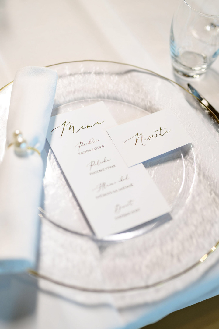 A close-up of a plate featuring a white menu, the name of the guest, and a white napkin tied with pearls. The plate is transparent with a golden rim.
