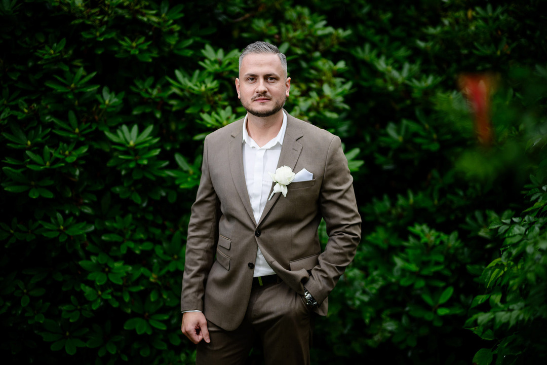 Photo of the groom alone in the garden, posing for the camera, smiling, standing confidently with one hand in his pocket.
