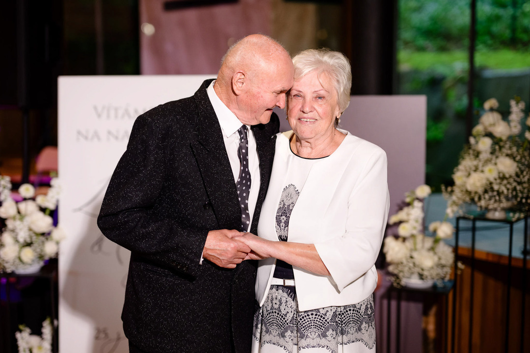 Grandparents pose together in a close embrace, holding hands, the lady looking and smiling directly at the camera, the gentleman leaning his face against his wife and smiling at her. A moment full of love and memories.
