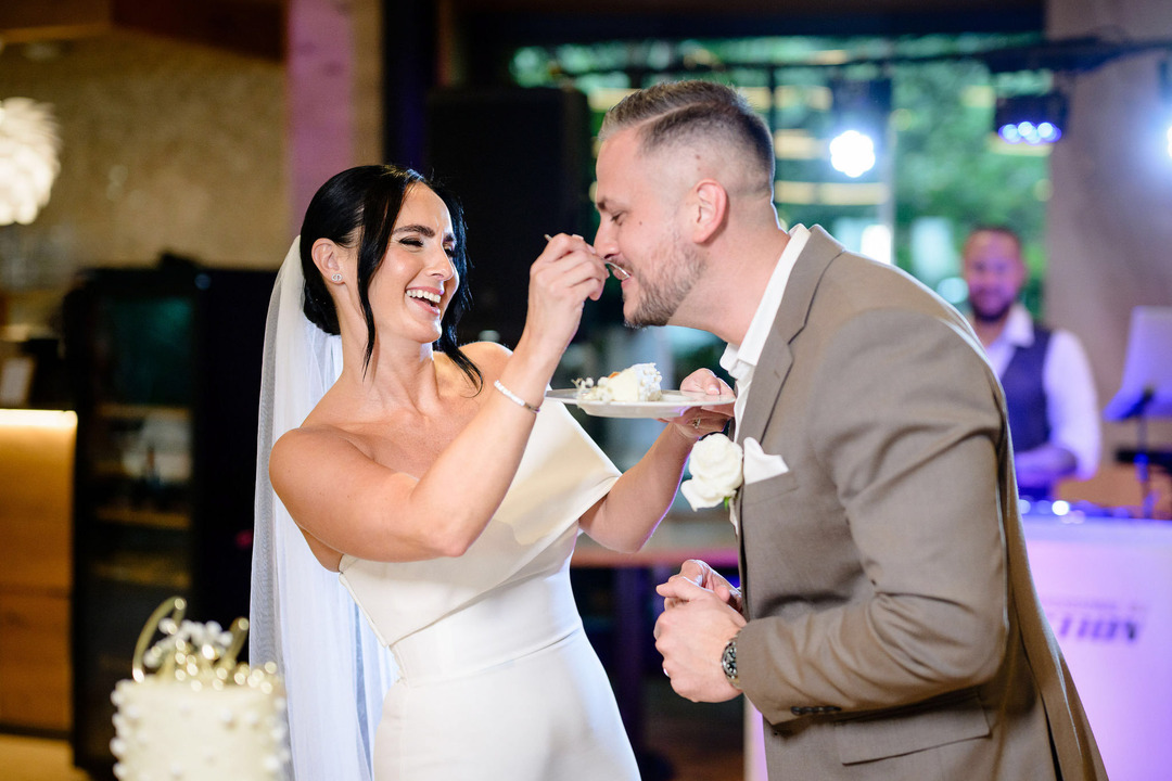 The bride feeds her new husband a piece of cake. They both laugh at the playful moment.
