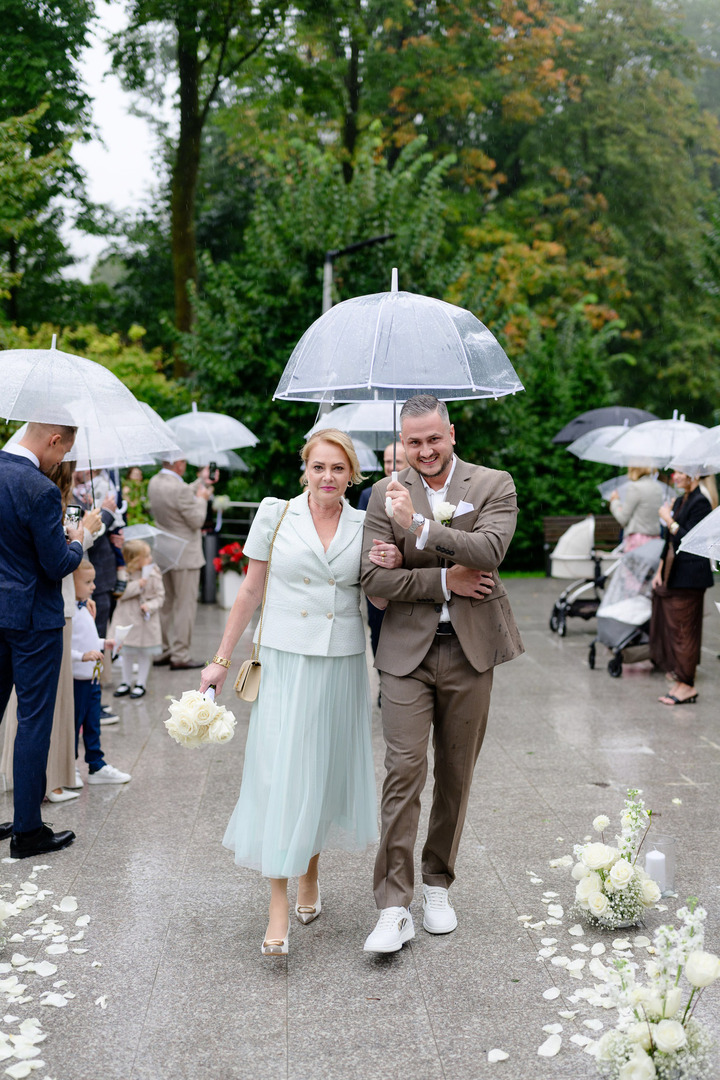 The groom walks down the aisle with his mother towards the wedding venue in the garden of Villa Cattaleya, holding an umbrella over both of them.
