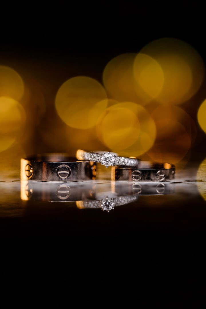 A close-up shot of Cartier wedding rings alongside an engagement ring, with the photograph having a darker tone. The rings are set against a bokeh of yellow lights in the background, creating a soft, romantic atmosphere.
