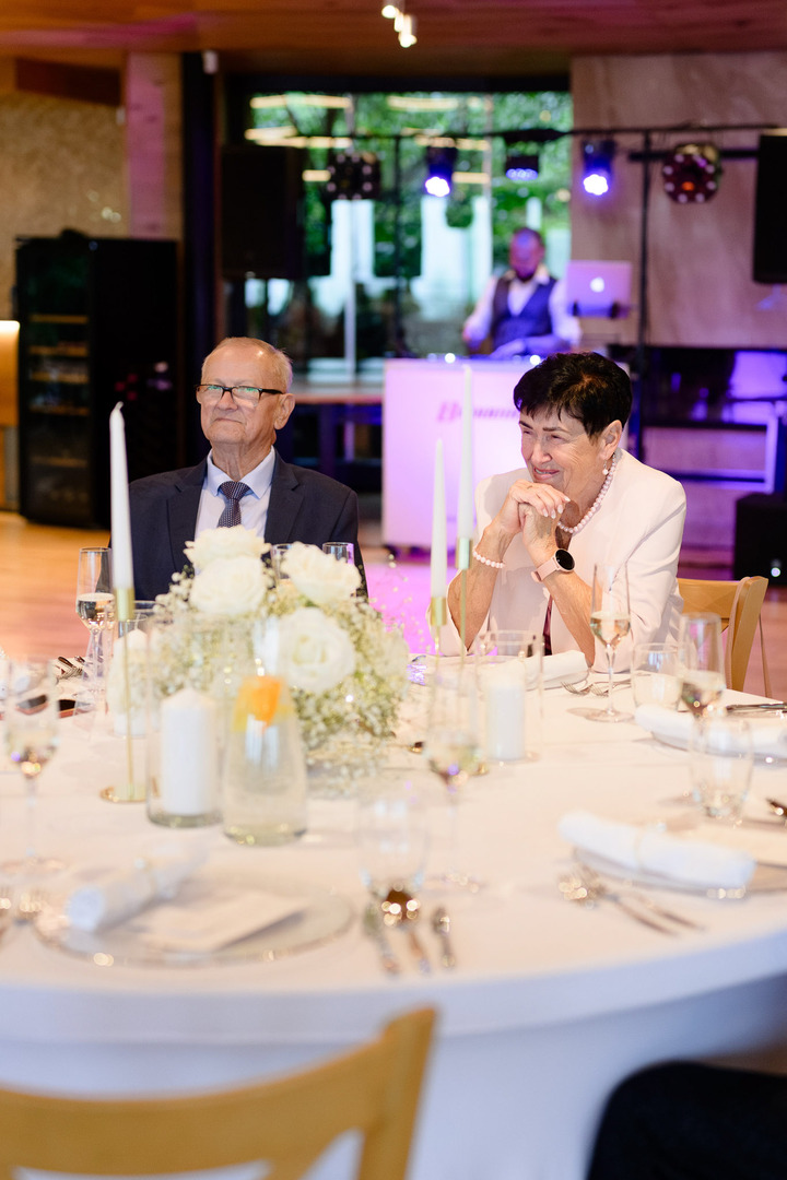 Grandparents sitting at a festively set table, listening to a speech and smiling.
