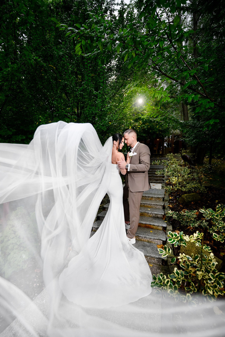 The newlyweds stand in a romantic moment in the garden of Villa Cattaleya in Čeladná. They are in close proximity, savoring a quiet moment amidst the busy day. The bride’s long veil gently flows toward the camera.