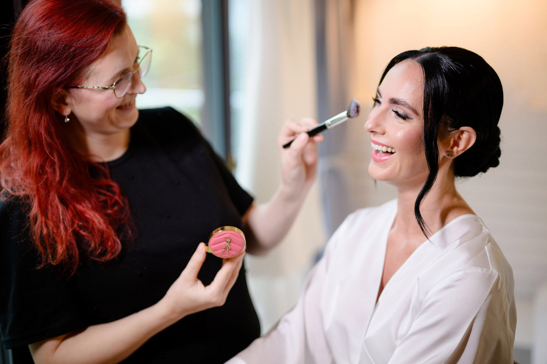 Makeup artist Petra Grezlova is applying makeup on the bride, and they are both laughing joyfully.
