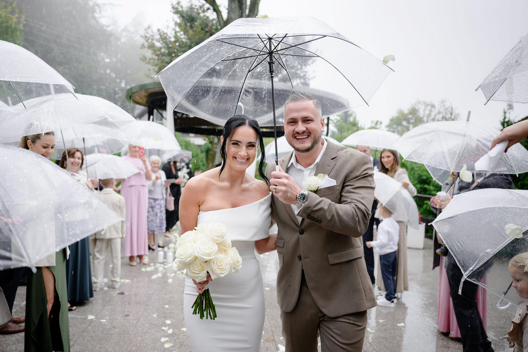 The bride and groom walk happily down the aisle, holding a transparent umbrella over them as it rains.
