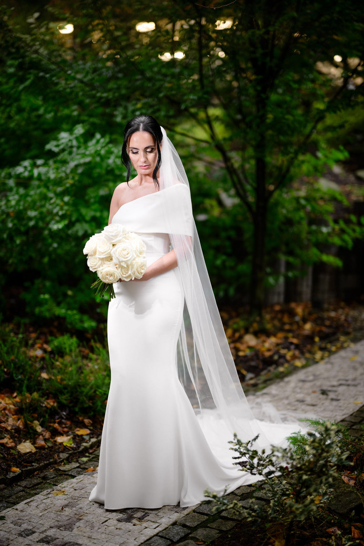 The bride, in elegant flowing dress with a long veil, stands in the garden of Villa Cattaleya, surrounded by lush greenery.
