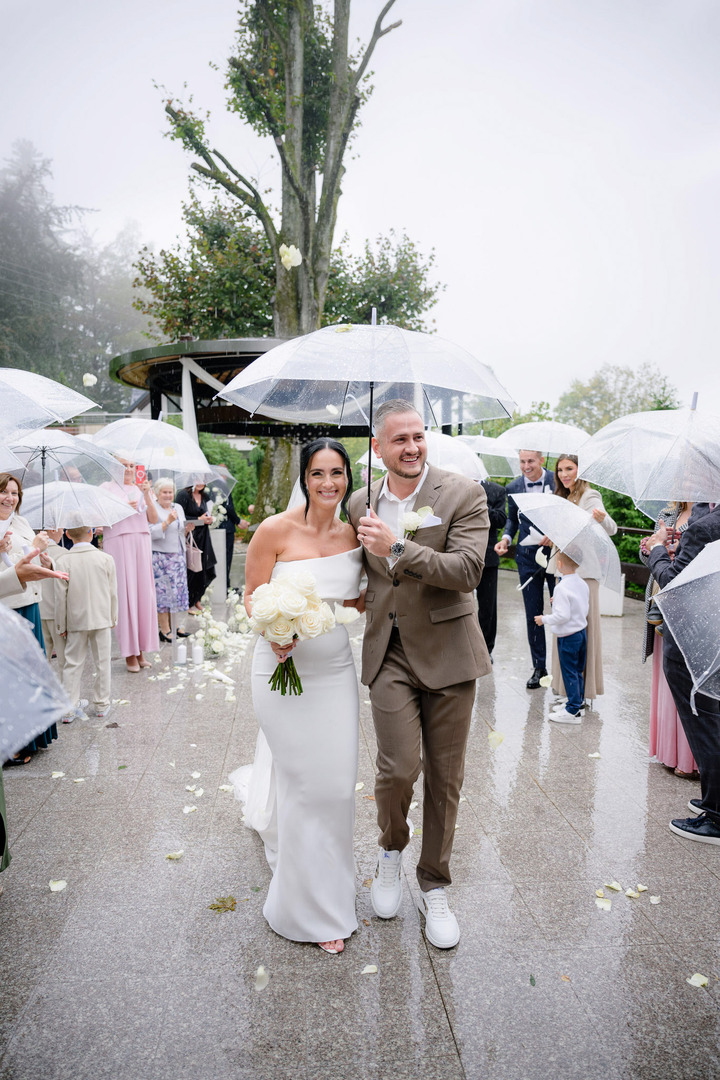 Newlyweds walking down the aisle of guests, holding an umbrella above them and smiling at the camera.
