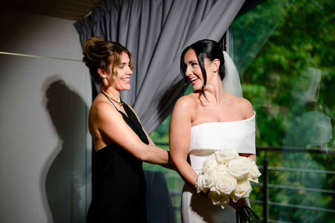 The maid of honor helps the bride into her white wedding dress. The bride holds a bouquet of white roses in her hand.
